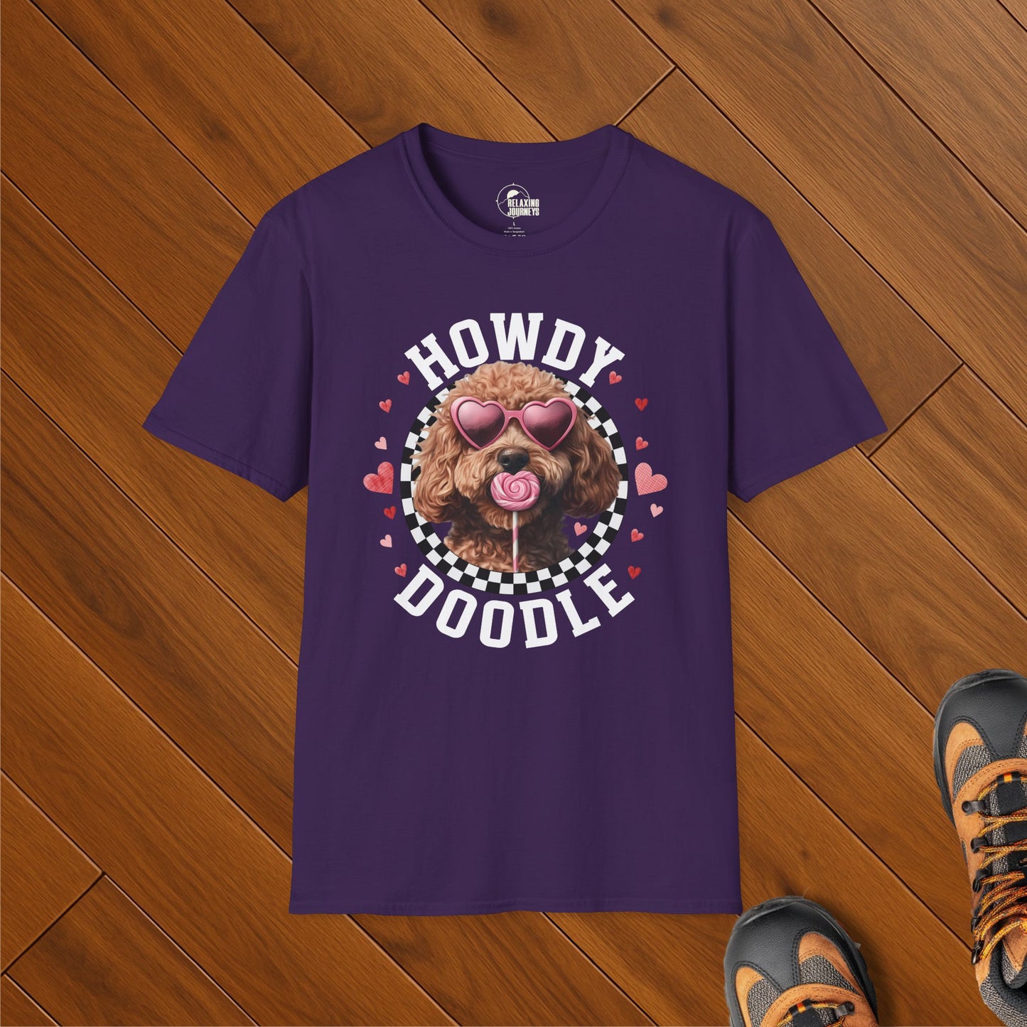 Purple t-shirt featuring a doodle dog wearing heart-shaped sunglasses and licking a lollipop, surrounded by a checkered circle and hearts with the text “Howdy Doodle.” Shirt is laid flat on wooden floor next to hiking boots.