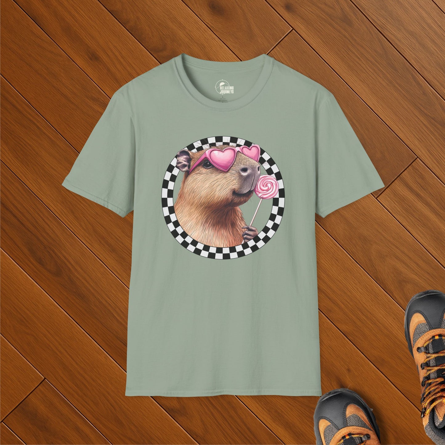 Sage green t-shirt featuring a smiling capybara wearing heart-shaped sunglasses and holding a pink lollipop, framed by a black-and-white checkerboard circle. Shirt is laid flat on wooden floor with hiking boots nearby.