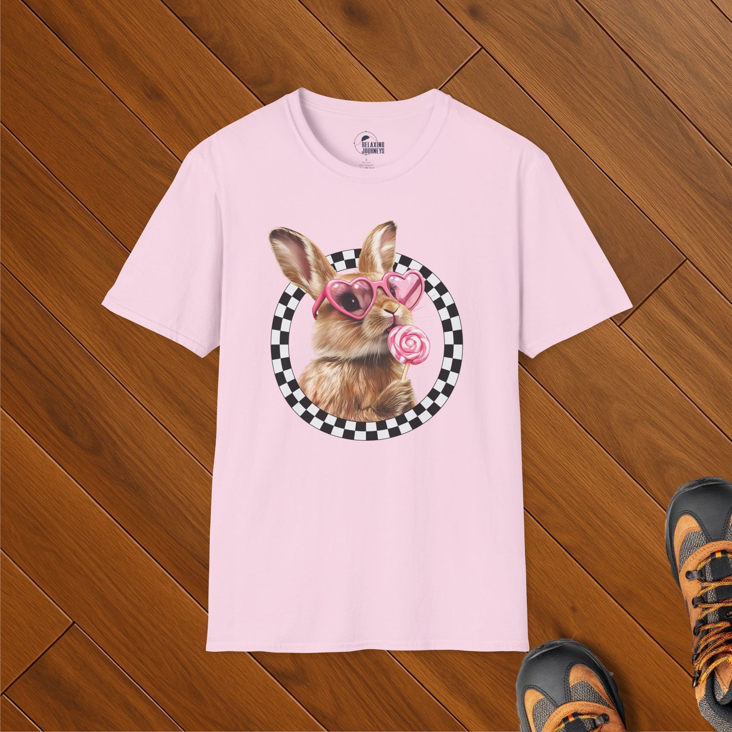 Light pink t-shirt featuring a cute bunny wearing pink heart-shaped sunglasses and holding a swirled lollipop, framed by a black-and-white checkerboard circle. Shirt is laid flat on wooden floor with hiking boots nearby.