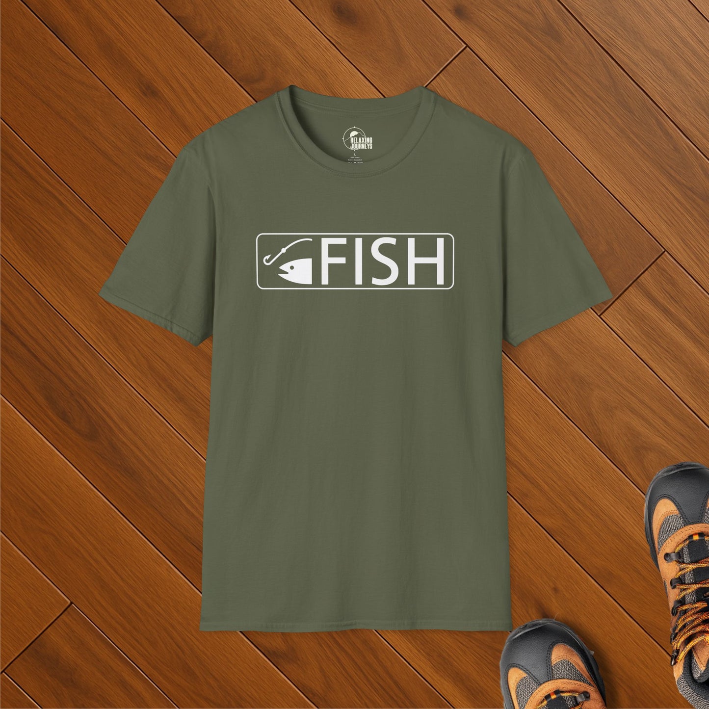 Olive green t-shirt with white FISH text and trail sign fishing hook icon, laid on wood floor with hiking boots