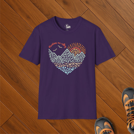Purple t-shirt with heart-shaped mountain sunrise design and “Mountains are calling” text, styled on wood floor with hiking shoes