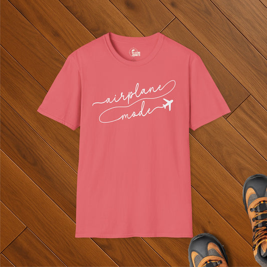 Coral pink t-shirt with white script text that reads “airplane mode” featuring a small airplane graphic. Shirt is laid flat on wooden floor with hiking shoes visible.