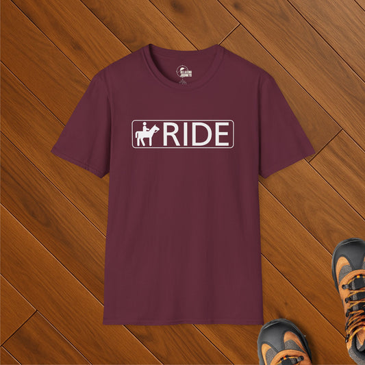 Maroon t-shirt with white RIDE sign featuring horseback trail icon, laid on wood floor with hiking boots