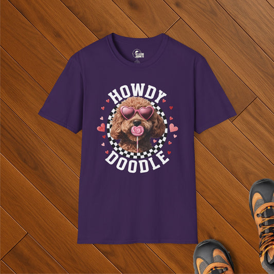 Purple t-shirt featuring a doodle dog wearing heart-shaped sunglasses and licking a lollipop, surrounded by a checkered circle and hearts with the text “Howdy Doodle.” Shirt is laid flat on wooden floor next to hiking boots.
