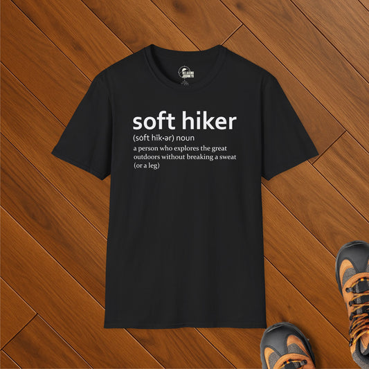 Black t-shirt with humorous "soft hiker" definition design, folded on wood floor with hiking shoes